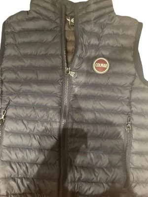Svart dunväst från Colmar - Snygg svart dunväst från Colmar med quiltad design ny kr 2000 och två dragkedjeförsedda fickor framtill. Västen har hög krage och Colmar-logga på bröstet. Perfekt för lager-på-lager och håller dig varm under kyliga dagar.