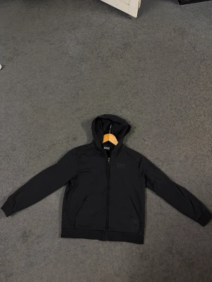 Svart hoodie från EA7 Emporio Armani - Säljer en svart hoodie från EA7 Emporio Armani med dragkedja framtill och två fickor. Klassisk huva och diskret logga på bröstet. Perfekt för en clean och stilren look. Materialet känns mjukt och bekvämt, passar till det mesta.