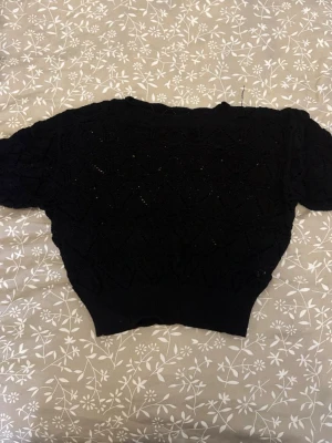 Svart stickad tröja med mönster - Säljer en svart stickad tröja med korta ärmar och snyggt hålmönster. Tröjan har ribbade muddar vid ärmslut och nederkant, vilket ger en skön passform. Perfekt för dig som gillar en enkel men ändå detaljerad look. Köpt i Spanien och har används 2-3 gånger och säljer då den är för liten och inte används.
