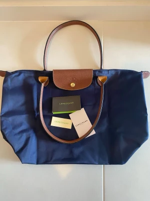 Mörkblå Longchamp Le Pliage väska - Stilren mörkblå handväska från Longchamp Le Pliage med bruna läderdetaljer och guldfärgad knapp. Väskan har klassisk form, vikbar design och två handtag i läder. Väskan är i nyskick. Använd 1 gång. 