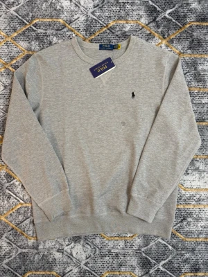 Grå stickad tröja från Polo Ralph Lauren - Snygg ljusgrå stickad tröja från Polo Ralph Lauren med klassisk rund halsringning och den ikoniska svarta logotypen broderad på bröstet. Tröjan har ribbade muddar vid ärmslut och nederkant. Perfekt för en clean och stilren look.