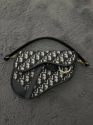 Svart och beige saddle handväska - Trendig saddle handväska i svart och beige med monogrammönster och guldfärgade metalldetaljer. Väskan har en unik form, kort axelrem och dekorativ D-detalj framtill. Materialet är en mix av textil och läderdetaljer. Perfekt accessoar för att lyfta din outfit.FEJK