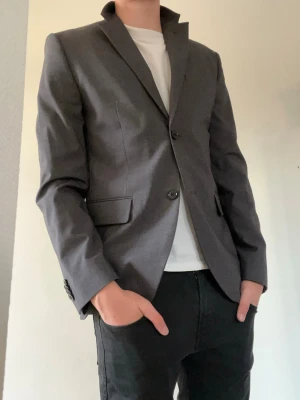 Mörkgrå klassisk kavaj - Tjena!👋 Säljer denna snygga mörkgrå kavaj med två knappar framtill och klassisk krage från H&M i storlek S🤩Kavajen har två fickor med lock och en bröstficka😮‍💨Passformen är rak och materialet ser ut att vara en blandning av polyester och viskos, vilket ger en stilren look😤 Meddela vid frågor!📩