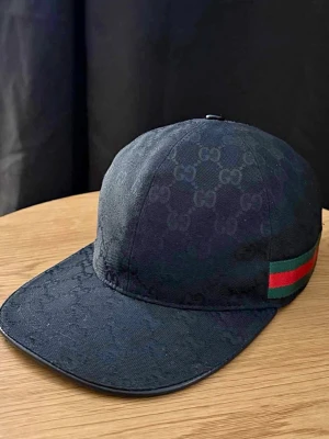 Svart Gucci keps  - Svart keps från Gucci med diskret GG-monogram över hela tyget och klassiska gröna och röda ränder på sidan. Justerbar rem baktill för perfekt passform. Snygg och stilren accessoar som lyfter din outfit.