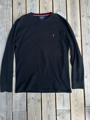 Svart långärmad tröja Polo Ralph Lauren - Svart långärmad tröja från Polo Ralph Lauren i 100% bomull. Tröjan har rund halsringning med röd kant och den klassiska röda logotypen broderad på bröstet. Enkel och stilren design som passar till det mesta.