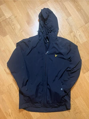 Svart vindjacka från Lyle & Scott - Säljer denna snygga svarta vindjacka från Lyle & Scott med dragkedja i storlek S (men kan även passa M), huva med dragsko och två fickor framtill. Jackan har det klassiska gula logobroderiet på bröstet och meshfoder på insidan. Perfekt för blåsiga dagar och enkel att matcha med olika outfits. Vid frågor, extra bilder eller liknande är det bara att kontakta mig!