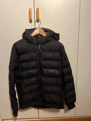 Svart pufferjacka från Nike med huva - Säljer en svart pufferjacka från Nike med huva och dragkedja framtill. Jackan har en klassisk quiltad design, två sidofickor och elastiska muddar vid ärmsluten. Perfekt för kalla vinterdagar och riktigt skön att bära. Storleken på jackan är XL i barnstorlek! 