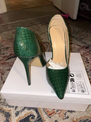 Gröna pumps med krokodilstruktur - Snygga gröna pumps med spetsig tå och hög klack. Skorna har en glansig yta med krokodilpräglat mönster och öppen insida. Perfekta för dig som vill sticka ut med ett par trendiga och eleganta klackar. OBS priset är inte hugget i sten ;)