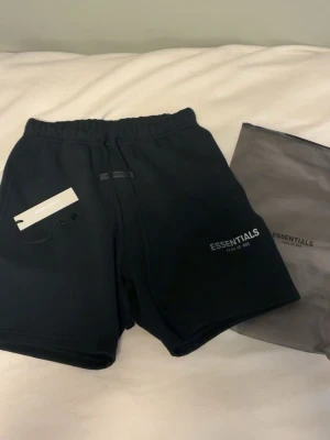 Svarta shorts från Essentials - Svarta shorts från Essentials med elastisk midja och snörning. Klassisk passform med tryckt logga på vänster ben och en bakficka. Perfekta för chill dagar eller till träningen. Kommer med originalpåse och tag kvar.