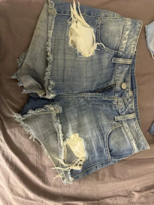 Guess shorts Blå slitna jeansshorts med fransar - Säljer ett par blå guess denimshorts med slitna detaljer och fransiga kanter. Shortsen har hög midja, dragkedja och knapp framtill samt klassiska fickor både fram och bak. Perfekta för sommaren och ger en avslappnad vibe.