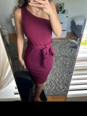 Vinröd oneshoulder klänning med knyt - Säljer en vinröd, figurnära oneshoulder klänning med knytband i midjan. Klänningen är kort och har en stilren, enkel design utan mönster. Perfekt för dig som vill ha en trendig och elegant look.