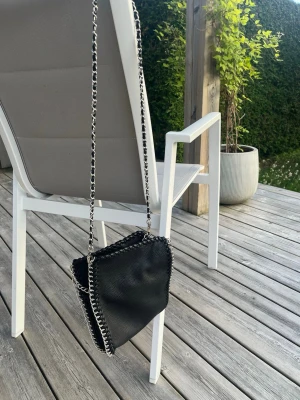 Svart axelväska med kedjerem - Snygg svart axelväska i konstläder med silverfärgad kedjerem och detaljer längs kanterna. Väskan har en mjuk, fyrkantig form och cool kedjesöm som ger en trendig look.