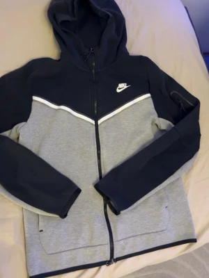 Nike hoodie i grått och svart - Nike hoodie med dragkedja i grått och svart, vit Nike-logga på bröstet och vit detalj över bröstet. Huva med dragsko och två fickor framtill. Perfekt för en sportig och avslappnad stil.
