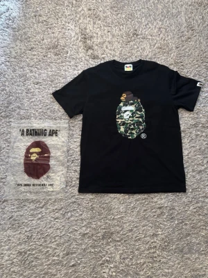 Svart T-shirt  - Säljer en svart t-shirt. Trycket har en grön camo-äggform och en liten apa ovanpå. Klassisk passform och rund hals. Perfekt för dig som gillar streetwear och vill sticka ut.