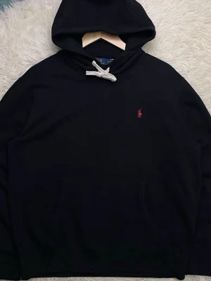 Svart hoodie från Polo Ralph Lauren - Klassisk svart hoodie från Polo Ralph Lauren med vit dragsko i huvan och röd broderad logga på bröstet. Hoodien har känguruficka framtill och är tillverkad i mjukt bomullsmaterial. Perfekt för en avslappnad och stilren look. Pris kan diskuteras!!