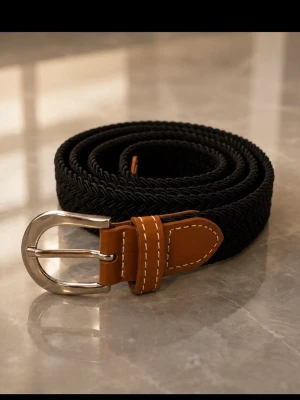 Svart flätat bälte med bruna detaljer - Snyggt svart flätat bälte med bruna skinnliknande detaljer och silverfärgat spänne. Perfekt accessoar för att lyfta din outfit med en klassisk touch. Bältet har vita sömmar som kontrast mot det bruna och svart flätad rem.