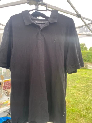 Svart pikétröja från Jack & Jones - Klassisk svart pikétröja från Jack & Jones med krage och knappar framtill. Tröjan har korta ärmar och är tillverkad i ett mjukt bomullsmaterial. Perfekt för en stilren och enkel look.