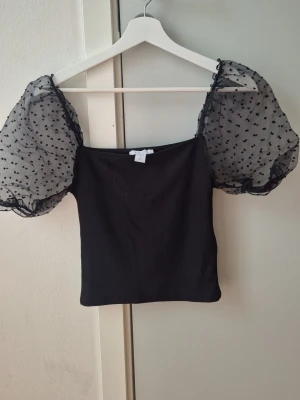 Svart topp med puffärmar  - Trendig svart topp med transparenta puffärmar i mesh dekorerade med små hjärtan. Toppen har en croppad passform och fyrkantig urringning, perfekt för att sticka ut lite extra. Ärmarna ger en romantisk och lekfull vibe.