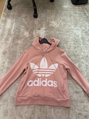 Rosa hoodie från Adidas med logga - Säljer en snygg rosa hoodie från Adidas med stor vit Trefoil-logga och text på bröstet. Hoodien har huva med dragsko och en avslappnad passform. Perfekt för dig som gillar streetwear och vill ha något bekvämt och stilrent.