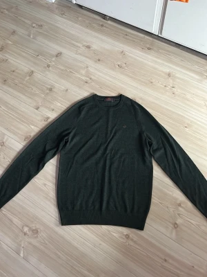 Mörkgrön stickad tröja från Morris - Snygg mörkgrön stickad tröja från Morris i 100% merinoull. Tröjan har rund halsringning, diskret logga broderad på bröstet och ribbade muddar vid ärmslut och nederkant. Perfekt för dig som gillar klassisk och stilren design.
