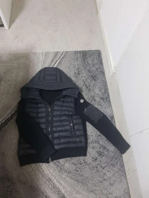 Moncler-cardigan - Snygg moncler cardiganjacka med quiltad kropp och stickade ärmar. Jackan har huva, dragkedja framtill och en ficka med dragkedja på bröstet. Ärmarna är ribbstickade och har en patchdetalj på vänster arm. Perfekt för lager-på-lager och streetwear.