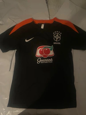 Svart Brasilien Nike fotbollströja - Svart kortärmad fotbollströja från Nike med Brasilien-logga och Guaraná Antarctica-tryck på bröstet. Snygga orange detaljer på axlarna och klassisk rund hals. Perfekt för dig som älskar fotboll och vill sticka ut på planen.