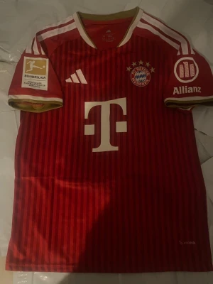 FC Bayern München matchtröja röd - Röd FC Bayern München matchtröja med vita och guldiga detaljer, klubbmärke och sponsorer på bröstet samt nummer 17 och namnet Olise på ryggen. Adidas-logga på framsidan och Bundesliga-märke på ärmen. Perfekt för fotbollsfans!
