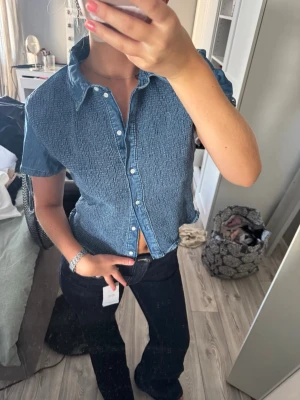 Kortärmad blå blus - Säljer en kortärmad skjorta i blå denim-look med klassisk krage och knappar framtill. Skjortan har en avslappnad passform, perfekt för en chill och stilren vibe. Passar till både jeans och kjol för en casual look. Köpte för ett tag sen men har aldrig använt. Nypris: 379 kr