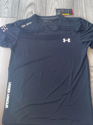 Svart träningsset från Under Armour - Snyggt svart träningsset från Under Armour med t-shirt och shorts. T-shirten har vita loggor och textdetaljer, rund hals och korta ärmar. Shortsen har elastisk midja och vita detaljer på sidorna. Perfekt för gymmet eller löpning.