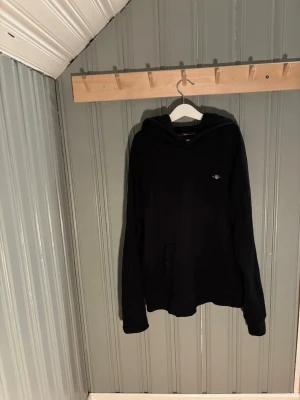 Svart hoodie med liten logga - En svart hoodie med huva och magficka. Liten diskret logga på bröstet. Klassisk passform med långa ärmar och ribbade muddar. Perfekt för en avslappnad stil. Materialet ser ut att vara mjukt och bekvämt, troligen bomull eller bomullsblandning.