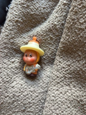 Söt figur med hatt och gitarr - En liten plastfigur föreställande en karaktär med stor gul hatt och orange kläder som håller i en silverfärgad gitarr. Figurer har stora ögon och ett leende ansikte. Perfekt som dekoration eller samlarpryl.