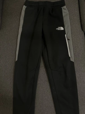 Svarta mjukisbyxor från The North Face - Snygga svarta mjukisbyxor från The North Face med gråa paneler längs sidorna. Byxorna har resår i midjan, dragkedjeficka på ena benet och logga både fram och bak. Perfekta för chill eller träning, tillverkade i mjukt material.