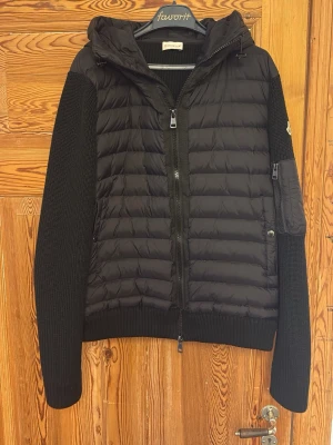 Svart moncler hybridcardigan - Stilren svart pufferjacka med quiltad front och huva. Ärmar och rygg är stickade för en schysst kontrast och extra komfort. Jackan har dragkedja framtill och en ficka med dragkedja på ärmen. Perfekt för kalla vinterdagar. Den är liten i storleken eftersom Moncler är kända för att gå ner i storlek. Så den sitter ungefär som en stor M eller L. Den har fungerande NFC och QR-kod.