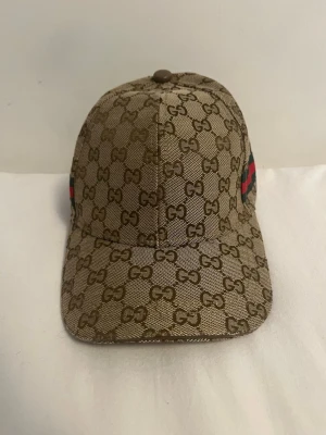 Beige keps med GG-mönster - Snygg beige keps med klassiskt GG-monogram över hela kepsen och en grön-röd rand på sidan. Kepsen har böjd skärm och är tillverkad i ett slitstarkt tygmaterial. Perfekt för dig som vill ha en exklusiv och trendig accessoar.