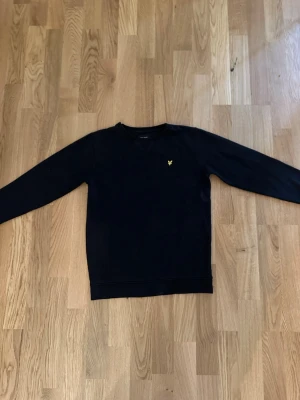 Lyle & scoot tröja - En svart stickad tröja med rund hals och lång ärm. På bröstet finns en liten gul broderad logga som detalj. Tröjan har ribbade muddar vid ärmslut och nederkant. Perfekt till höst och vinter för en clean look.