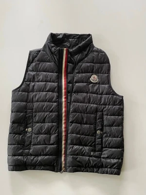 Moncler väst - Snygg svart dunväst med quiltad design och hög krage. Västen har två fickor med tryckknappar och en dragkedja framtill med röd, vit och blå rand. Moncler-logga på bröstet ger en exklusiv touch. Perfekt för lager-på-lager och Grisch och streetstyle. Har en väldigt liten laglapp baktill se bild nr 7