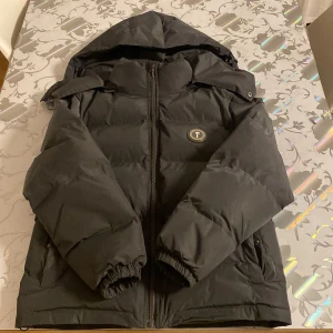 Grå pufferjacka från Trapstar - Snygg grå pufferjacka från Trapstar med broderad text på ryggen och ett runt märke på bröstet. Jackan har en dragkedja framtill och en stor huva för extra värme. Perfekt för kyliga dagar! Inga problem med jackan exakt som ny, knappast använd. Luvan är dock sönder men den går och sy ihop kostar typ 50-100kr