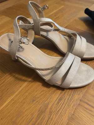 Beige pumps med remmar och strass - Snygga beige pumps med smala remmar och dekorativ strassdetalj. Skorna har öppen tå, justerbart spänne vid ankeln och en låg klack. Materialet ser ut att vara mockaimitation och de passar perfekt till en dressad look.