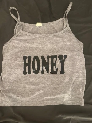 Grå linnetopp med texten HONEY - Säljer en grå linnetopp med tunna axelband och trycket 'HONEY' i svart framtill. Toppen har en croppad passform och är perfekt för dig som gillar en enkel men statement look.
