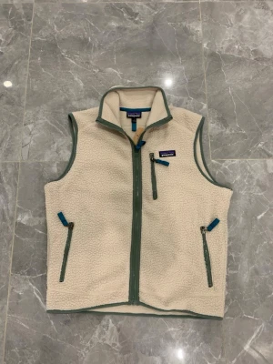 Beige fleeceväst från Patagonia - Säljer en beige fleeceväst från Patagonia med dragkedja framtill och tre fickor med blå detaljer. Västen har hög krage och är gjord i mjukt, värmande material. Perfekt för lager-på-lager och outdoor-stil.