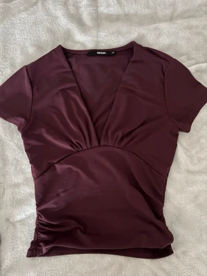 Vinröd v-ringad topp - Snygg vinröd topp från Bik Bok med djup v-ringning och korta ärmar. Modellen är figurnära och har en lätt rynkning i midjan. 