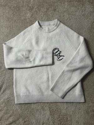 Ljusgrå stickad tröja med brodyr - Ljusgrå stickad tröja med rund halsringning och ribbade muddar. På bröstet finns en svart broderad text med bokstäverna 'OKY' i snirklig stil. Tröjan har en avslappnad passform och är perfekt för kyligare dagar.