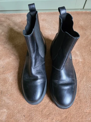 Svarta Chelsea boots i skinn - Snygga svarta Tommy Hilfiger Chelsea boots i äkta skinn med klassisk rund tå och elastiska paneler på sidorna. Grov sula ger en cool look och gör dem perfekta för höst och vinter. Lätta att dra på tack vare dragflikar både fram och bak. Använda några gånger. 