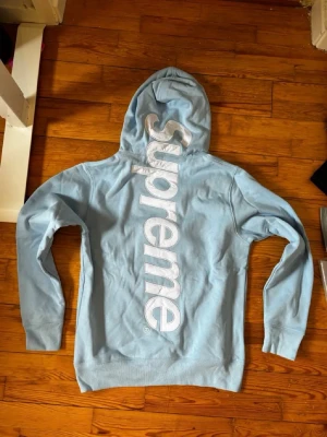 Ljusblå Supreme hoodie med stor logga - Supreme hoodie i ljusblått med stor vit Supreme-logga vertikalt på ryggen och klassisk röd Supreme-tag i halsen. Hoodien har huva med dragsko och en stor magficka framtill. Perfekt för dig som vill sticka ut med streetwear-vibe.