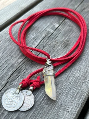 Rött halsband med kristall och mynt - Unikt halsband med rött mockaband, ett hängsmycke i form av en slipad kristall infattad i silverfärgad tråd samt två dekorativa metallmynt. Perfekt för dig som gillar boho-stil och vill ha något som sticker ut.