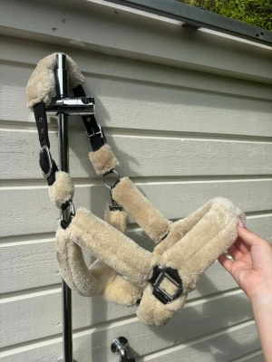 Beige fluffig grimma  - Säljer denna grimma med mysig, mjuk, fluffig vaddering och svarta detaljer. Grimman har metallspännen och ringar för enkel justering och fastsättning. Storlek full/storhäst 