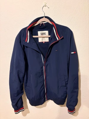 Marinblå vindjacka Tommy Jeans - Snygg marinblå vindjacka från Tommy Jeans med dragkedja, ribbade muddar och krage med röd, vit och blå detalj. Jackan har två sidofickor och diskret logga på ärmen. Tillverkad i lätt och tåligt material, perfekt för blåsiga dagar. M men passar hyfsat bra till S💫