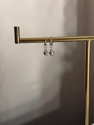 Små silverfärgade hjärtörhängen - Säljer ett par stilrena örhängen i silverfärg med små hjärtformade hängen. Örhängena har en enkel och modern design som passar till många olika stilar. Materialet ser ut att vara metall med blank finish. Perfekta för dig som gillar minimalistiska smycken.