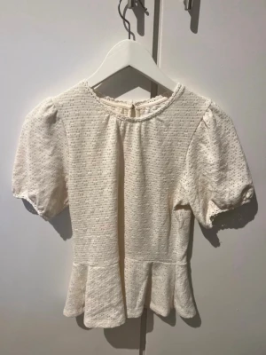 Vit blus med puffärm och volang - Söt vit blus med puffärmar och volang nedtill. Blusen har rund halsringning och knäppning bak i nacken. 