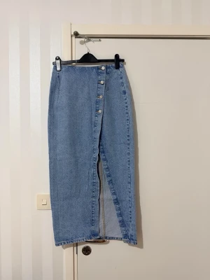 Lång jeanskjol med knappar och slits - Säljer en ljusblå lång jeanskjol med hög midja, knappar framtill och en snygg slits. Kjolen har två bakfickor och klassisk denimlook. Perfekt för dig som gillar stilrena och trendiga plagg.